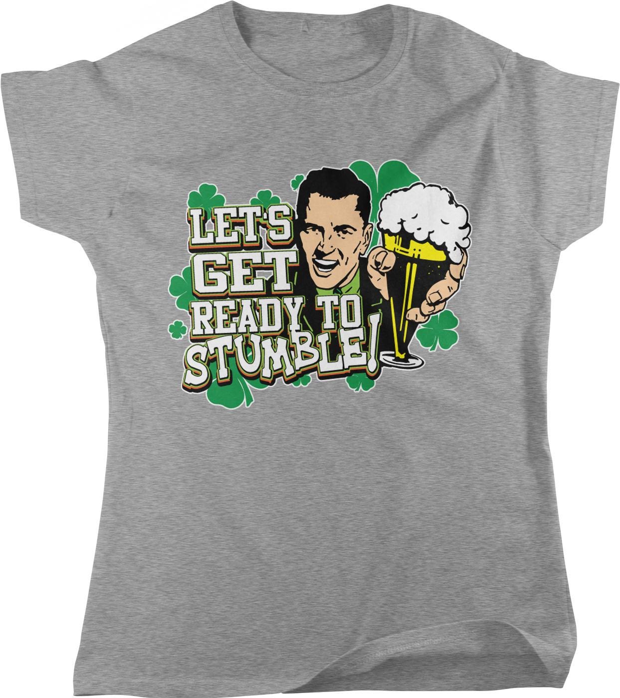 A gray t-shirt with a graphic design featuring a man holding a beer glass and a cloverleaf pattern. The text on the t-shirt reads "Let's Get Ready to Stumble!"