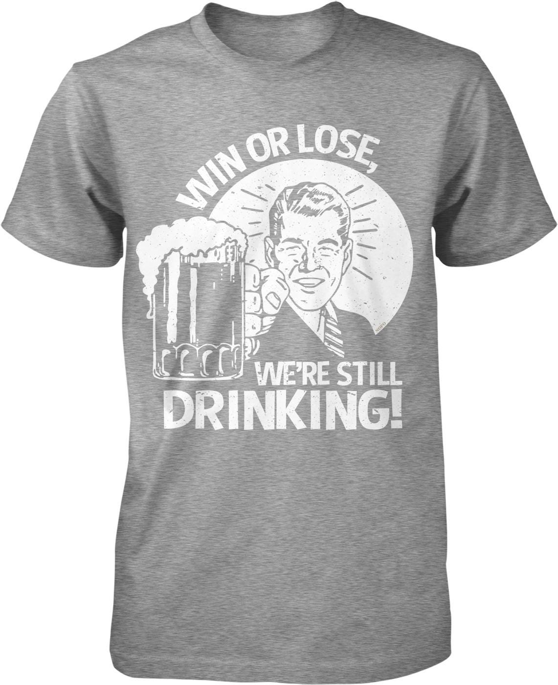A gray t-shirt with a graphic design depicting a man holding a beer and the text "WIN OR LOSE WE'RE STILL DRINKING!"