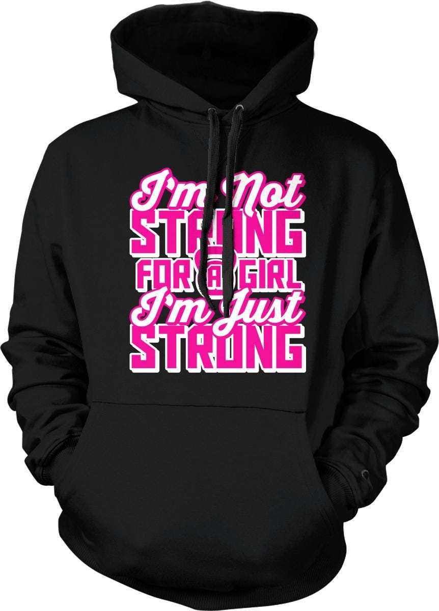 A black hoodie with a pink and white text design that reads "I'm not strong for a girl, I'm just strong".